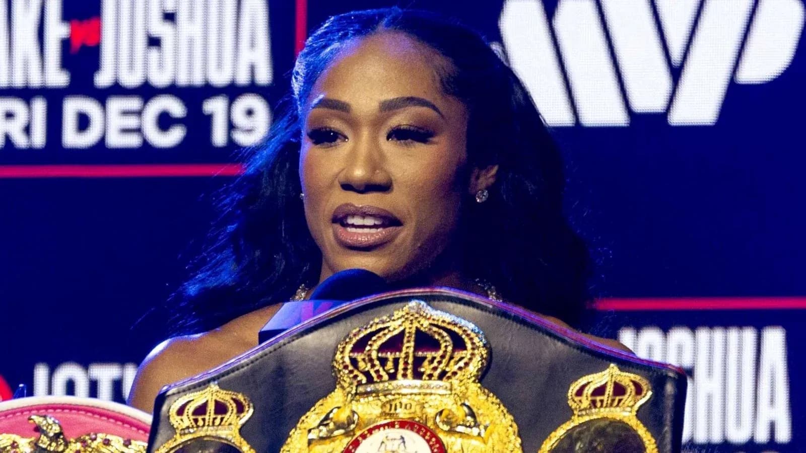 A woman speaking with boxing belts at podium
